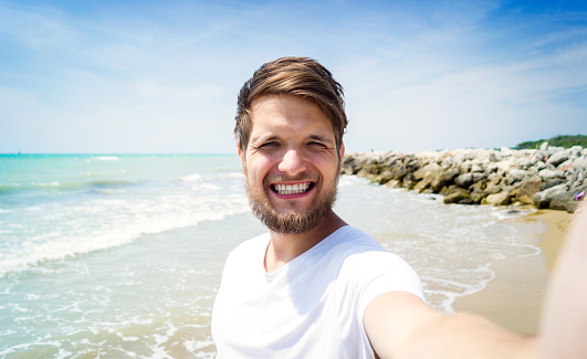 Handsome hipster man in white t-shirt on beach, smiling, taking selfie. Enjoying time at seaside.