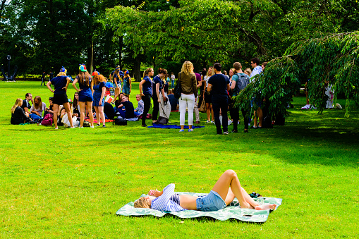 Lund, Sweden - August 24, 2016: Young adult woman lying on a blanket in a public park while students prepare for student orientation week in the distance.
