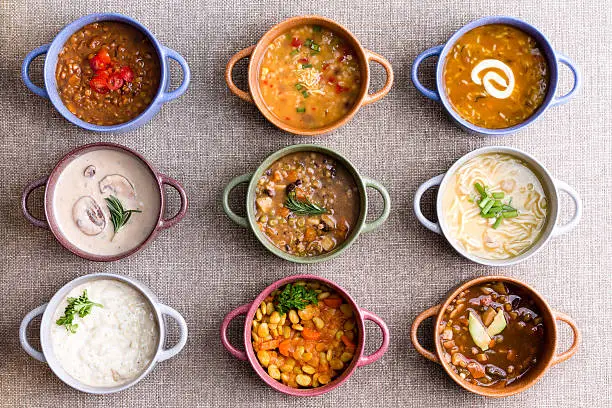 Assorted soups from worldwide cuisines displayed in bowls in three colorful lines garnished with cream and herbs in a World Of Soup concept, overhead view