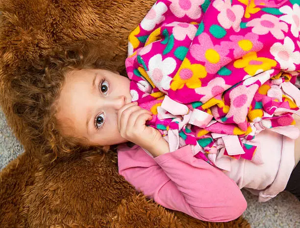 Young girl laying on the floor on top of a large brown stuffed animal. She is holding her pink floral security blanket and sucking her thumb as she looks up at the camera.