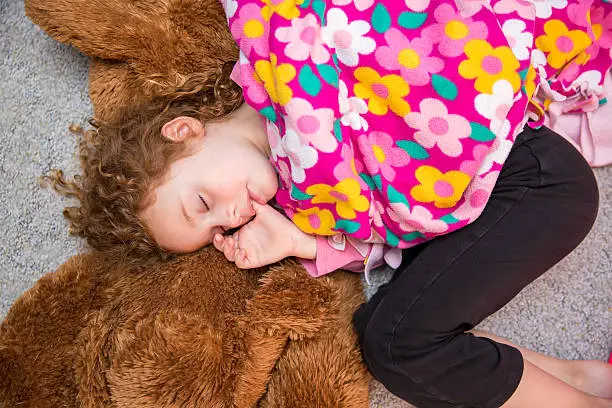 Young girl laying on the floor on top of a large brown stuffed animal. She is sucking her thumb and holding her pink floral security blanket.