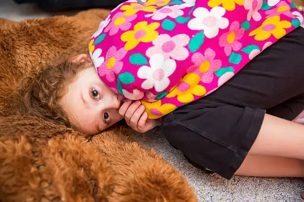 Young girl laying on the floor next to a large brown stuffed animal. She is holding her pink floral blanket around her and sucking her thumb as she looks at the camera.
