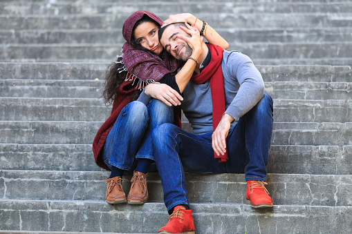 Young couple having fun, sitting on stairs. Both wears warm clothes. Woman with scarf on head, embracing the man. Stone stairs on background.