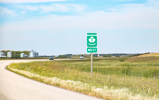 Horizontal image of the divided Trans-Canada Highway with green and white highway sign.  The road curves to the left and then the right with grain bins visible on the flat prairie landscape on the western edge of the province of Manitoba.