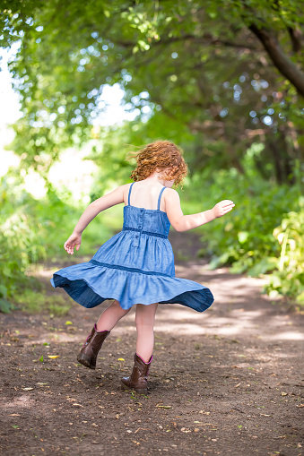 Rear view of a young girl twirling in circles on an outdoor dirt trail through the woods. The girl has curly red hair and is wearing a blue denim dress with brown cowgirl boots on this summer day on the farm.