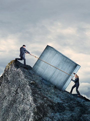 A businessman and a businesswoman attempt to move a large concrete block up a steep rocky mountaintop.  The businessman uses a rope to pull as the businesswoman tries to push from below.