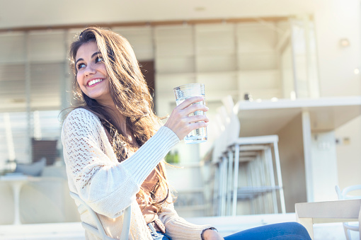 Woman drinking a glass of water. She is sitting on the terrace outside her luxury home. Kitchen in the background. She is smiling. Copy space