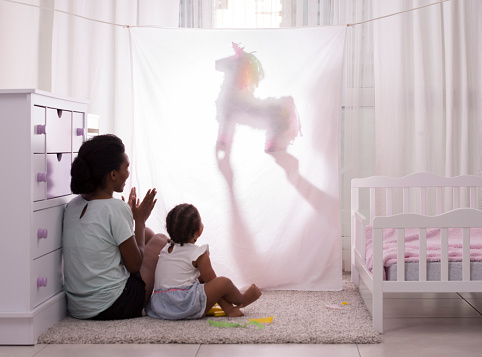 Father performing puppet shadow show to his 2.5 years old daughter and wife. Actor dimming the light with a unicorn puppet toy behind white curtain. Curious child enjoying to watch. Mother applauding.