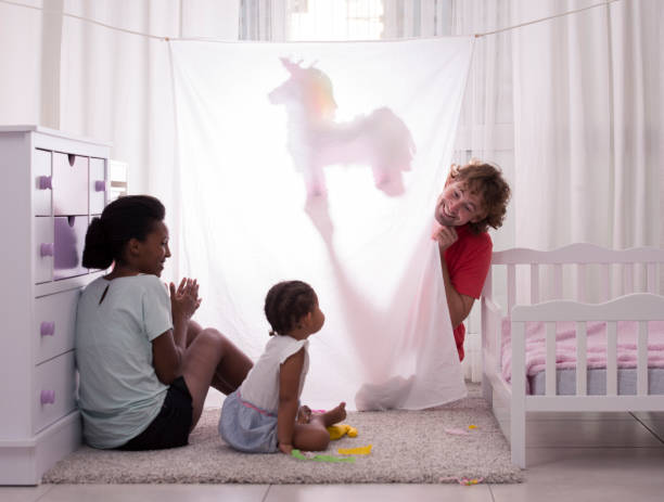 Father performing puppet shadow show to his 2.5 years old daughter and wife. Actor dimming the light with a unicorn puppet toy behind white curtain.