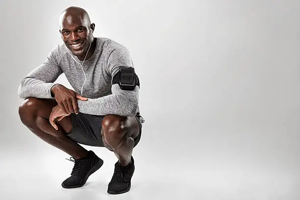 Smiling young african man with mobile phone armband and headphones crouching on grey background with copy space. Black male model listening to music.