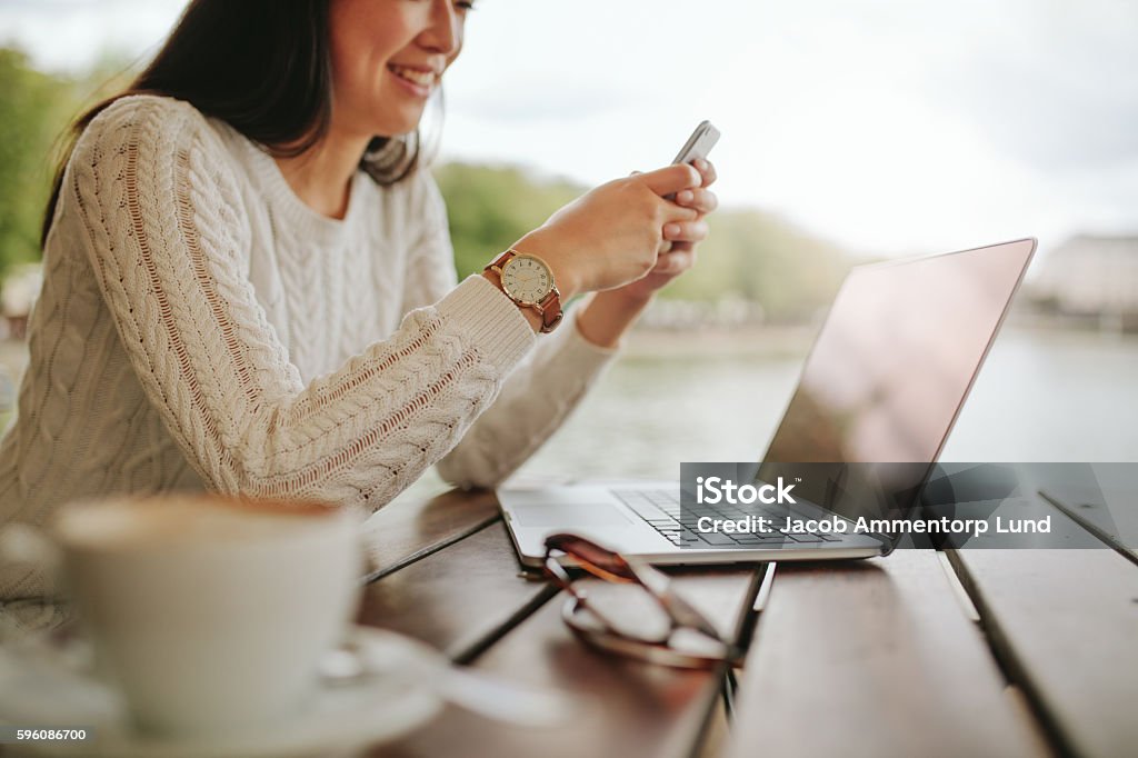 Young woman using mobile phone at outdoor cafe Cropped shot of young woman using mobile phone at outdoor cafe. Female sitting at table with laptop reading text message on her cellphone. Outdoors Stock Photo