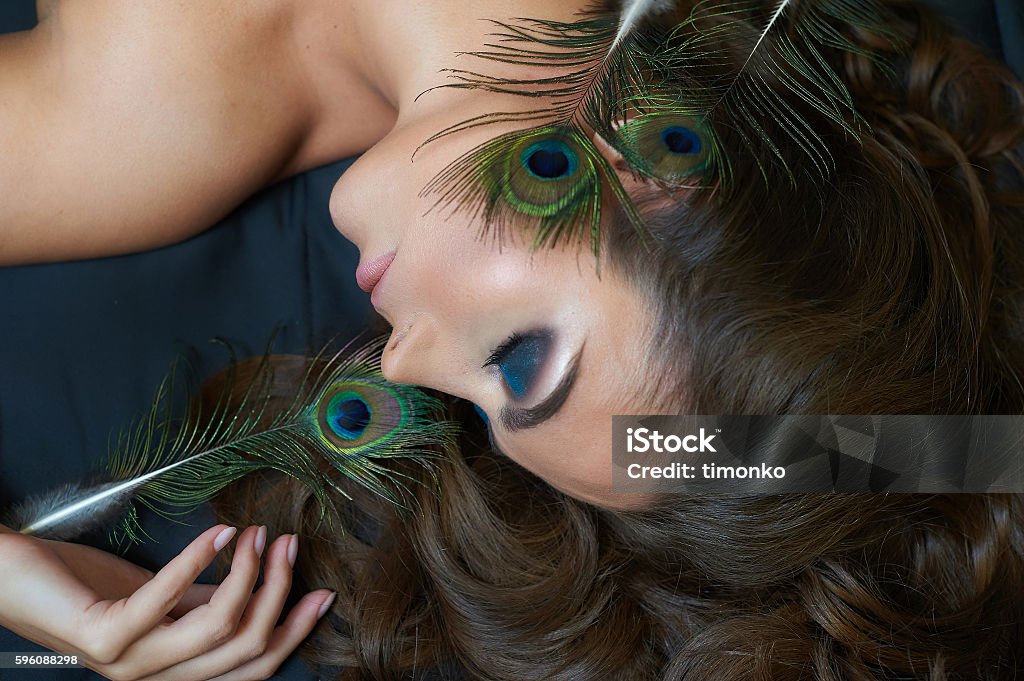 beautiful woman lying on bed with peacock feather and makeup beautiful woman lying on a bed with a peacock feather and makeup. Adult Stock Photo