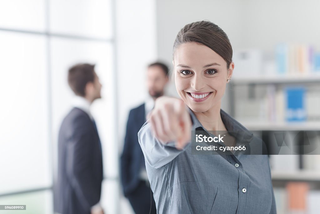 Smiling businesswoman pointing at camera Smiling businesswoman in the office pointing at camera, office interior and business people in the background, recruitment and career concept Adult Stock Photo