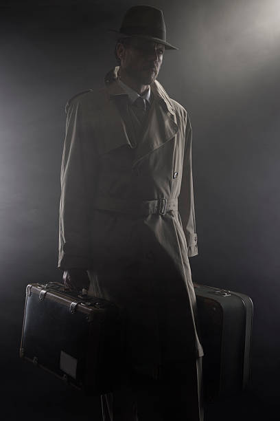 Man in trench coat with hat and luggage leaving in the dark, 1950s style film noir