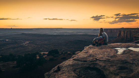 Father and son sitting on the edge of cliff overlooking vast landscape at Grand View Point of Canyonlands National Park during sunset.