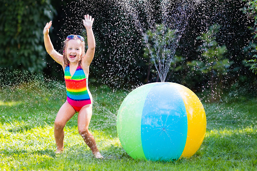 Funny laughing little girl in a colorful swimming suit playing with toy ball garden sprinkler with water splashes having fun in the backyard on a sunny hot summer vacation day