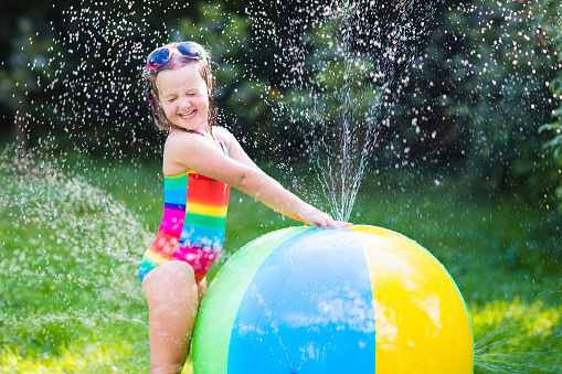 Funny laughing little girl in a colorful swimming suit playing with toy ball garden sprinkler with water splashes having fun in the backyard on a sunny hot summer vacation day