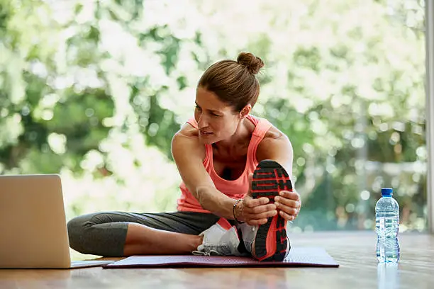 Full length of fit woman looking at laptop while doing stretching exercise at gym