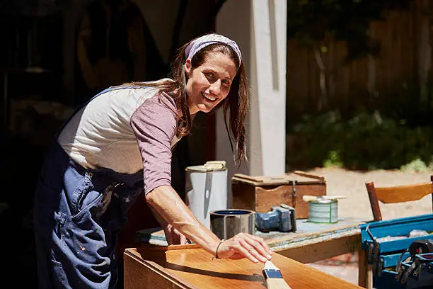 Portrait of happy mid adult woman applying varnish wooden furniture outside house
