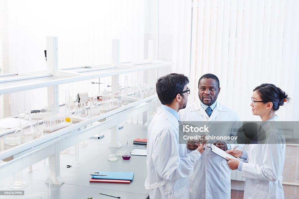 Scientific Discussion in Laboratory High angle shot of pleased male African-American, male Latin-American and female Asian laboratory scientists in lab coats and glasses discussing scientific research. Laboratory Stock Photo