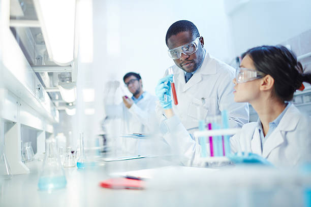Conducting Experiment in Laboratory Female Asian laboratory scientist in lab coat and safety goggles showing test tube with red liquid to curious African-American colleague in laboratory. Latin-American scientist in background. african american scientist stock pictures, royalty-free photos & images