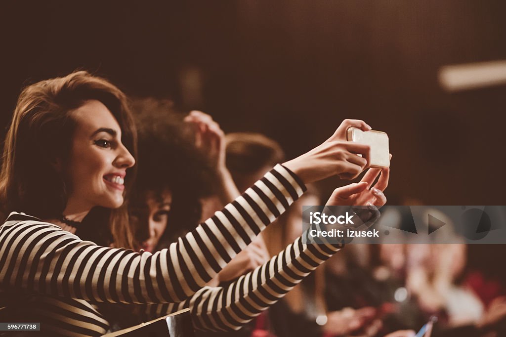 Young people at the concert, taking selfie using mobile Young people at the concert, teenager girl taking selfie using smart phone. Dark tone. Movie Theater Stock Photo