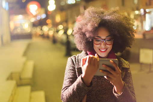 Beautiful young african woman with red eyeglasses and winter coat using digital tablet and walking on street at night. Buildings and lights as background.