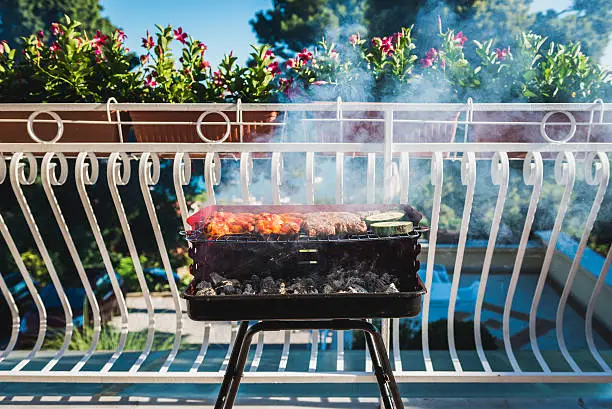 Delicious meat on barbecue grill with coal on balcony. Grilling food on a balcony or a terrace with wonderful view. Small cheap BBQ grill at home.