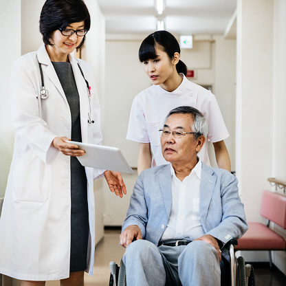 A female japanese doctor showing data on Digital Tablet to senior patient in a wheelchair. A nurse behind him pushing the chair.