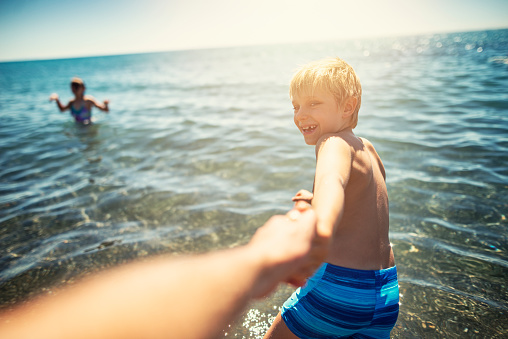 Little boy is pulling his father into the tuscan sea to play with him. Shot from father's personal perspective. The boy's sister is visible in the background. The boy is aged 7 and is laughing happily.