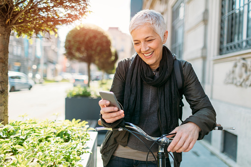 Mature woman enjoy her bicycle ride in the city. Cycling to and from her office. She just stopped to use her smart phone. Reading message or texting. Selective focus to her smiling face.