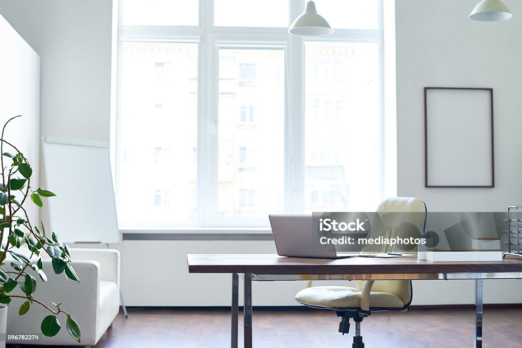 Empty Office Space Shot of empty modern well-lit office space with beige colored office chair and wooden desk with laptop on it standing before big window. Office Stock Photo