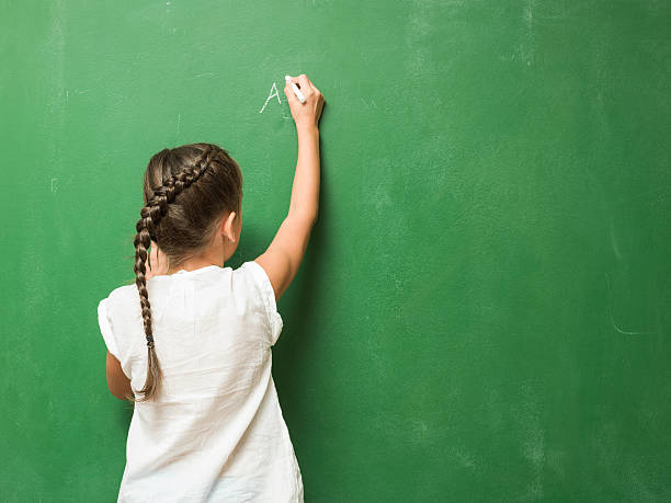 little girl writing on green chalkboard - child thinking writing little girls imagens e fotografias de stock