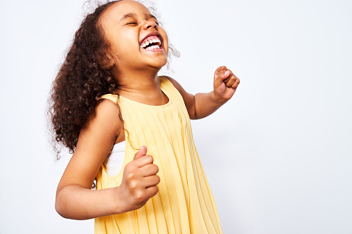 Portrait of African-American little curly haired girl in yellow dress laughing and dancing with closed eyes against white background