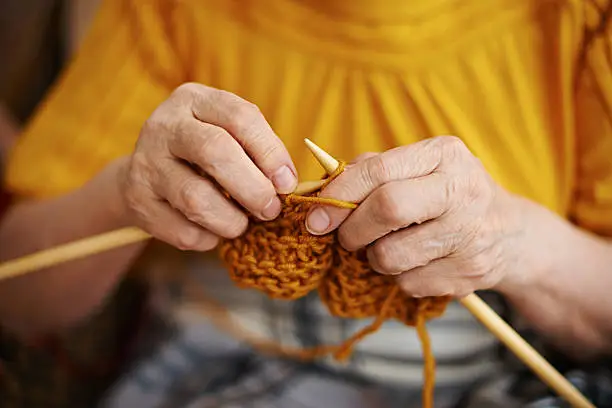 Close-up with selective focus of senior wrinkled female hands knitting with mustard colored yarn
