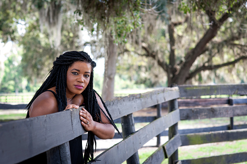 Beautiful black woman in 20-30s with long braided hair looks at camera while leaning on a fence on a horse farm with live oaks in the background in the country on a summer day, Marion County, Ocala, Florida, USA