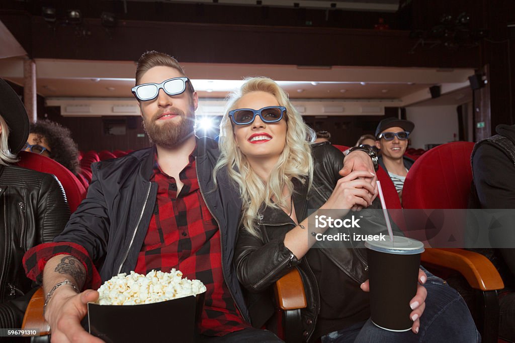 Bearded man and blonde woman in the 3D movie theater Group of young people in the 3D movie theater, wearing 3d glasses, watching comedy. Close up of blonde young woman and her bearded boyfriend holding popcorn. 3-D Glasses Stock Photo