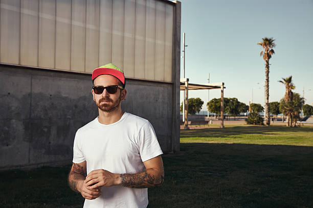 Tattooed young man wearing blank white t-shirt, trucker cap and sunglasses with a paper coffee cup on the background of concrete building and palms