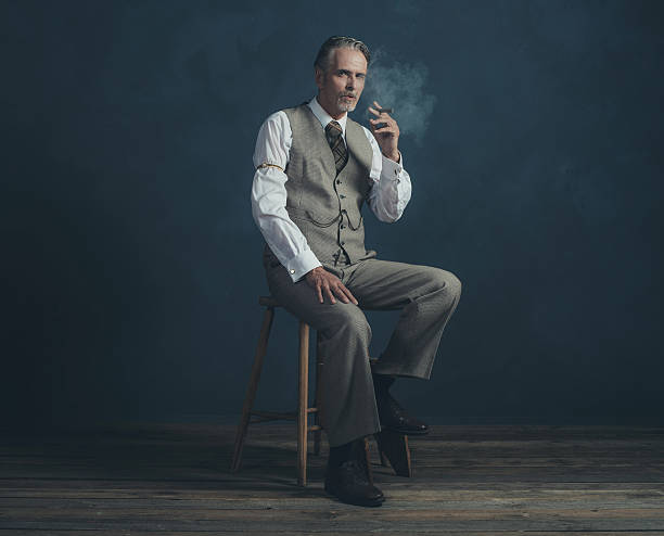 Cigar smoking retro 1920s man sitting on wooden stool. Cigar smoking retro 1920s man wearing suit sitting on wooden stool. Empty room. Vintage wooden floor. man cigar stock pictures, royalty-free photos & images