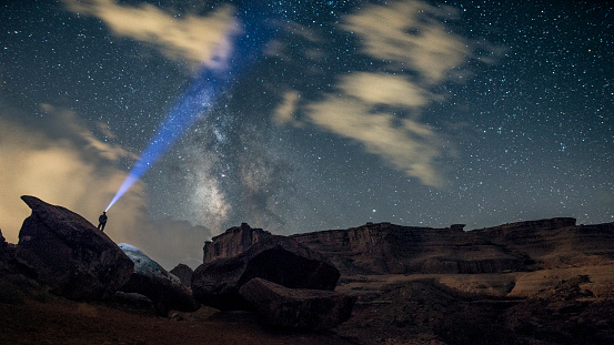 Man standing on the top of rock looking up with a headlight pointing toward Milky Way in Arches National Park, Utah, US on partly cloudy summer night.