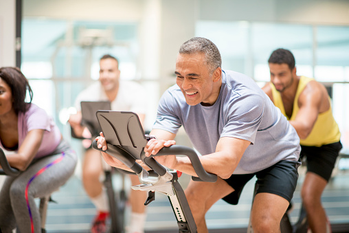 A multi-ethnic group of adults are taking a exercise class together at the gym. They are working by cycling on stationary bikes.