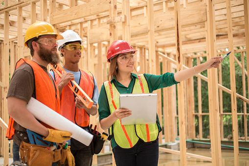 Female, Latin descent engineer, foreperson, or quality control inspector discusses blueprint plans with workers inside a construction job site.  The multi-ethnic team is working inside an unfinished framed house or small commercial building.  They are wearing safety vests, glasses, and hard hats.