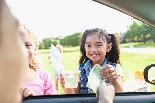 Pretty elementary age little girls gives a customer change after the customer purchases lemonade from the girl's lemonade stand. The customer is in the car and the girl hands the drink and change through the window.
