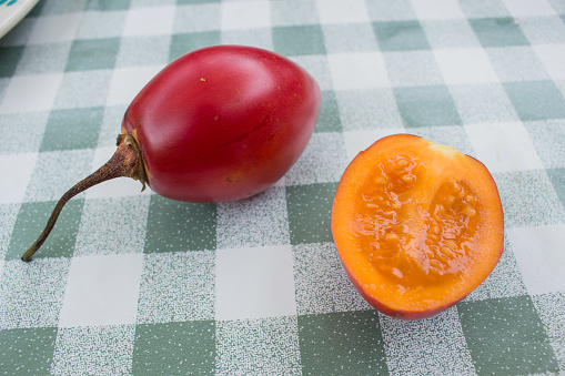 The healthy and exotic fruit tomate de arbol native to Colombia and Ecuador is lying on the table with checked cloth There is one uncut piece with red skin and another half of the fruit with visible yellow flesh. 