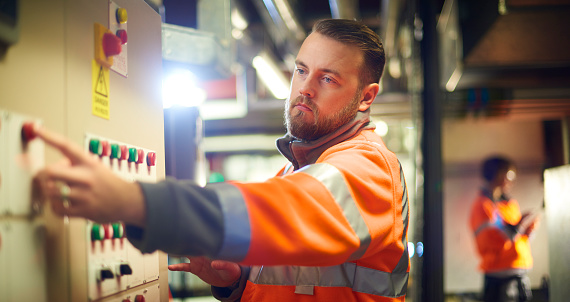 An industrial service engineer conducts a safety check of a control panel in a boiler room. He is wearing hi vis and is pressing one of the buttons on the control panel.