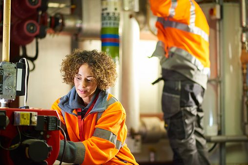 A female industrial service engineer and her colleague are in the boiler room of the plant facility that she works at. They are wearing hi vis and are conducting their regular safety inspection of one of the boiler room pumps.