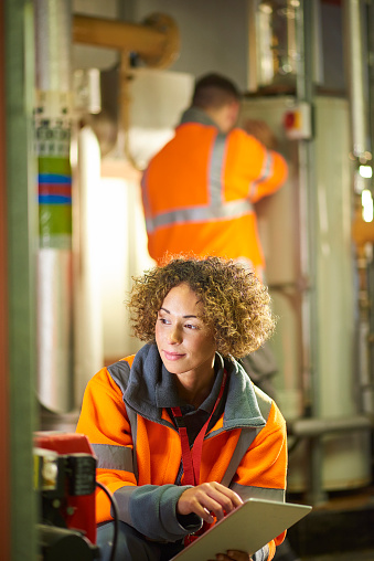 A female industrial service engineer and her colleague are in the boiler room of the plant facility that she works at. They are wearing hi vis and are conducting their regular safety inspection of one of the boiler room pumps.