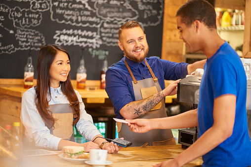 A female barista smiles as she holds the card machine for her customer to pay. The customer is paying with his card using the contactless method. A male barista is at the coffee machine just behind her preparing more customers drinks and having a chat with his colleague and the customer. The male barista is also the owner of the coffee shop.