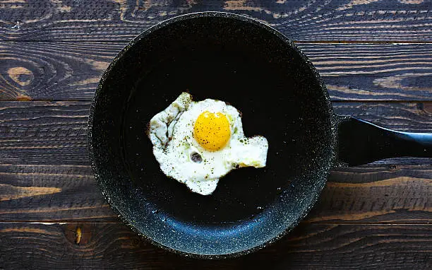 Fried egg in a black pan over a dark old dirty wood background with dramatic light.