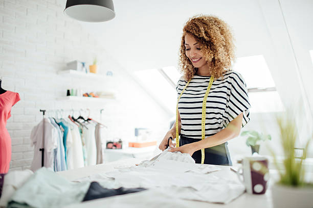 Latina Tailor In Her Workshop Latina tailor or fashion designer in her workshop. Standing by the desk and cutting fabric. Wearing tailor tape around neck. woman stitching stock pictures, royalty-free photos & images